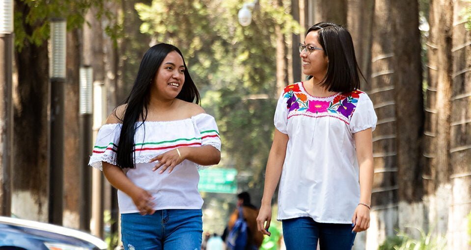 Two young Mexican women, walking on a sidewalk on a tree-lined street